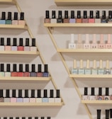 A neatly arranged display of nail polish bottles on triangular wooden shelves, featuring a variety of colors such as pastels, bright shades, and darker tones. The bottles are organized by color and brand, creating a visually appealing pattern.