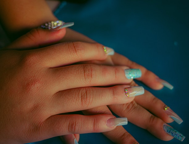 Hands with long, manicured nails displaying various nail art designs including rhinestones and glitter. The nails have a glossy finish, with some featuring sparkly turquoise and others clear with embellishments. The skin tone is warm and contrasts with the blue background.